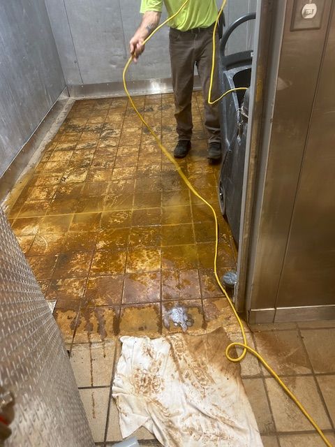 Person cleaning a dirty, stained tile floor with a machine and cord in a commercial kitchen area.