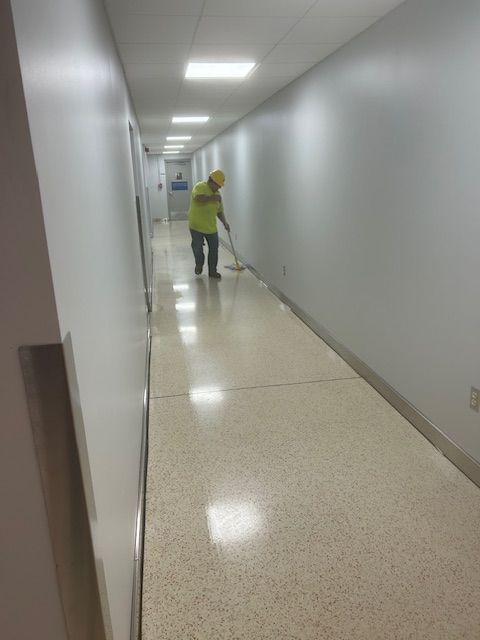 Person mopping a hallway with a yellow vest and hard hat. The hallway has white walls and a speckled floor.