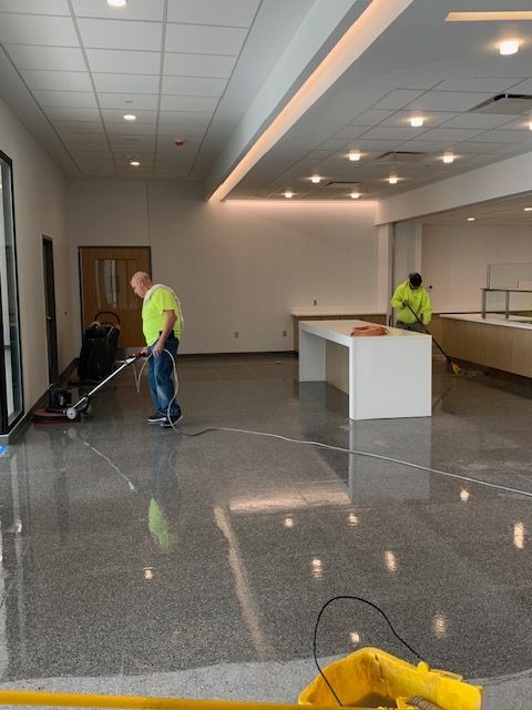 Two workers polishing a concrete floor in a room with white walls and a long white table.
