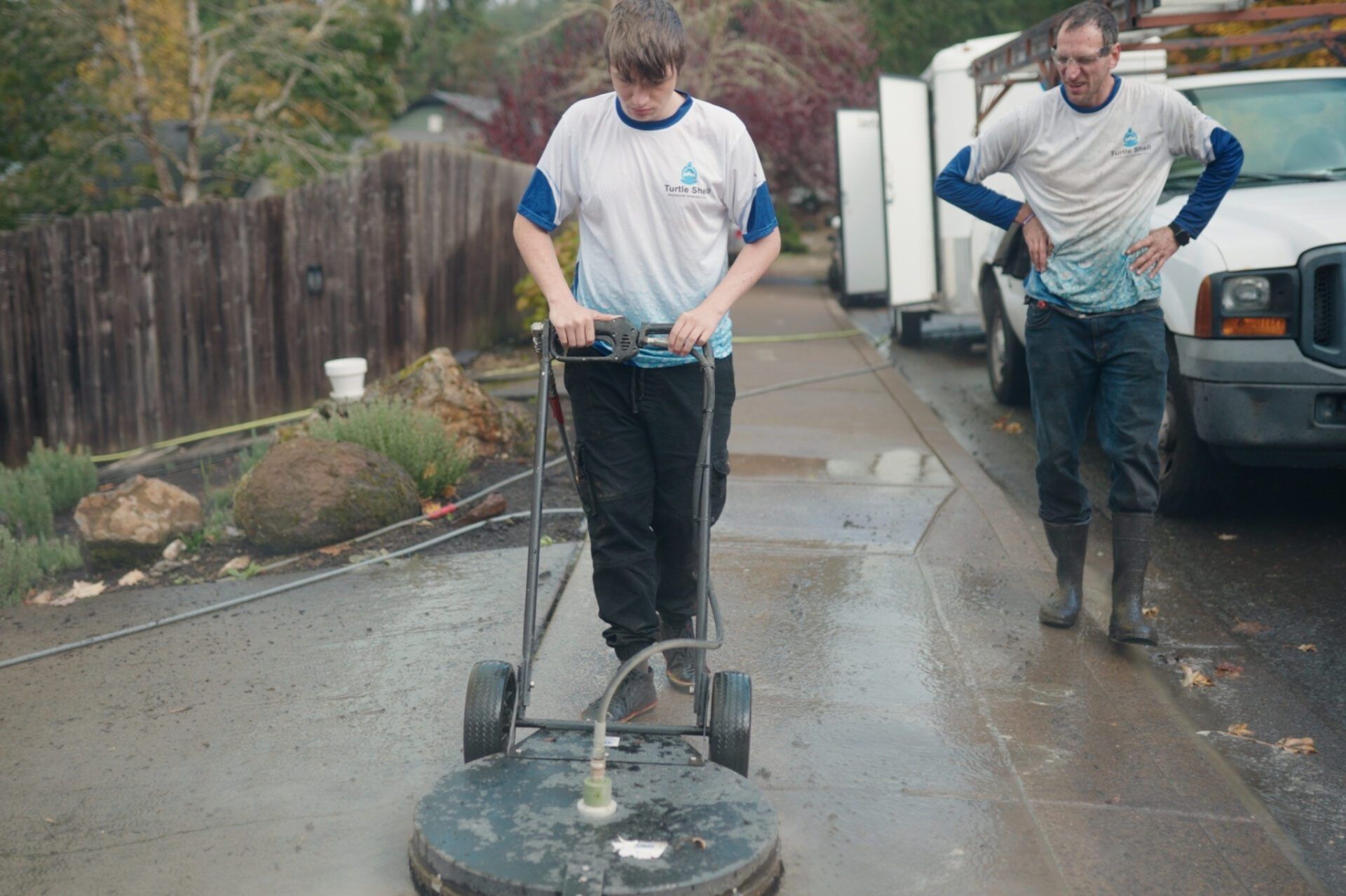Two workers pressure washing a wet sidewalk beside a house and parked car