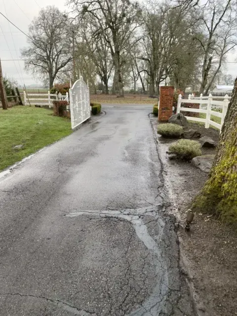 Wet driveway through open white gate, with brick pillars, trimmed hedges, and leafless trees.