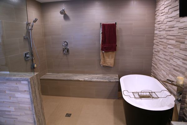 Modern walk-in shower with beige tile, built-in bench, handheld shower, red towel, and white tub on the right
