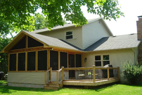 A beige house with a screened porch, wooden deck, and green lawn under leafy trees.