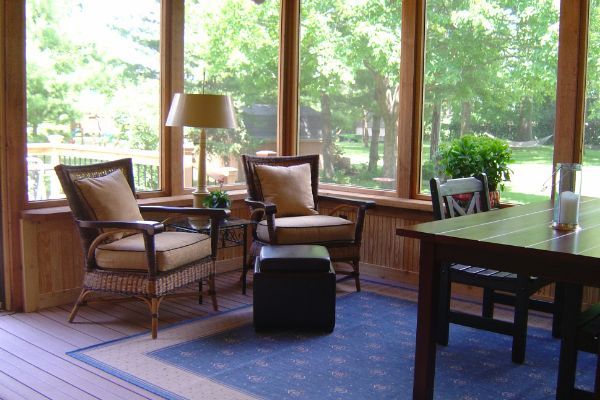 Wood-paneled sunroom with wicker chairs, lamp, rug, and a dark wooden table overlooking green trees.