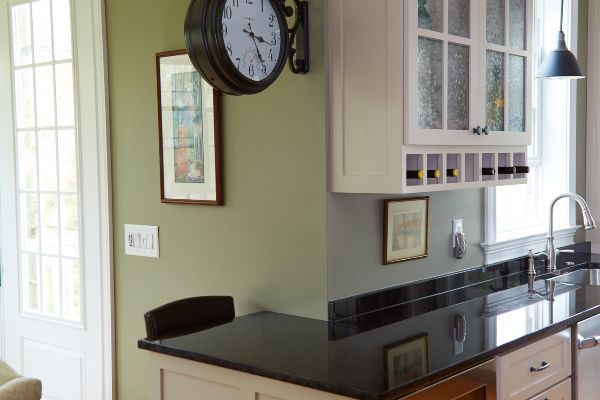 Modern kitchen with black countertops, white cabinets, wall clock, and stainless sink by a window.
