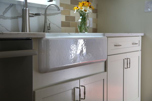 White kitchen sink with chrome faucet, glossy apron front, white cabinets, and a vase of yellow flowers.