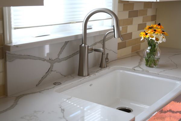Bright white kitchen sink with stainless faucet, marble backsplash, and a vase of yellow flowers by the window