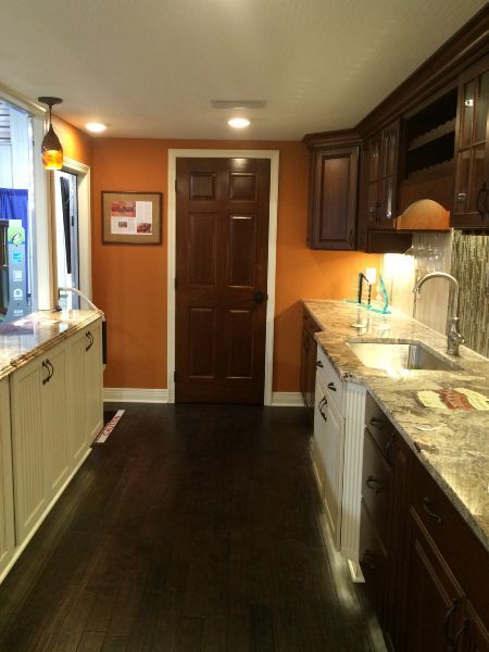 Narrow kitchen with dark wood floor, orange walls, granite counters, white cabinets, and closed wooden door.