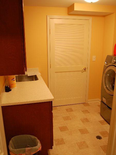 Small kitchen with white louvered door, beige tile floor, countertop, trash can, and stacked washer-dryer.