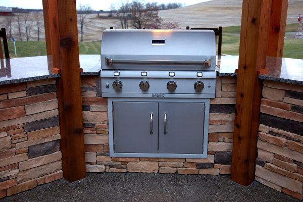 Stainless steel outdoor grill built into a stone counter under a wooden pavilion.