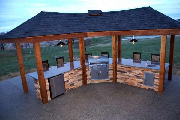 Wooden outdoor pavilion with built-in grill, cabinets, and hanging lights at dusk