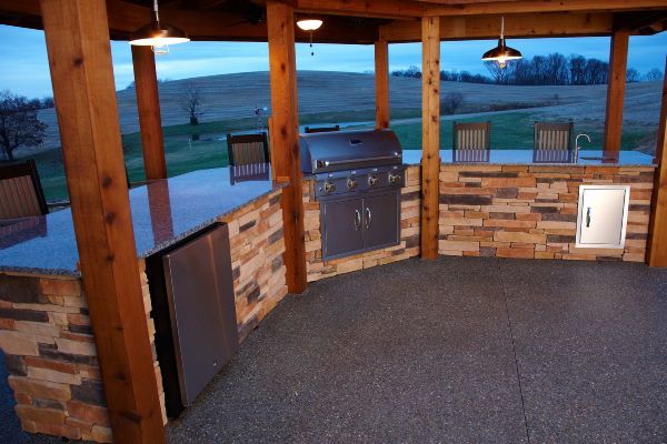 Covered outdoor patio with wood posts, stone wall, grills, and chairs overlooking a green landscape