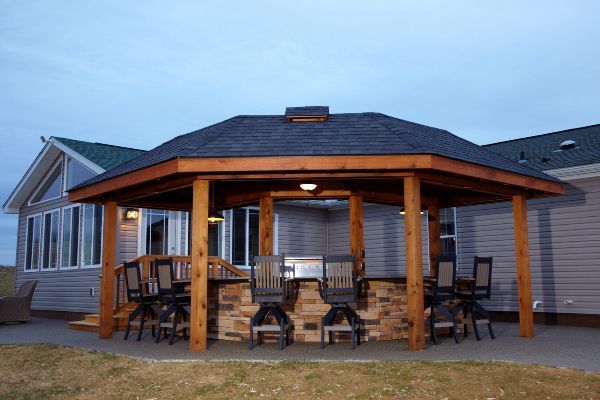 Covered outdoor patio with wooden beams, stone base, chairs, and a grill at dusk.