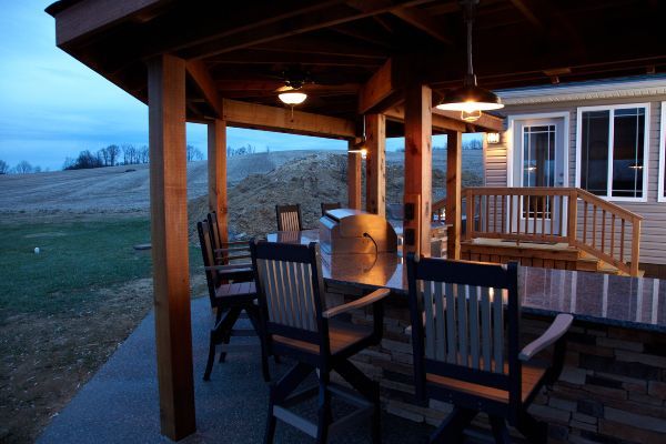 Covered porch with wooden chairs and table at dusk, overlooking open fields and a house exterior