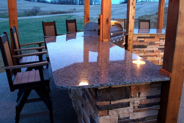 Covered outdoor patio with stone bar counter, wooden stools, and view of fields at dusk
