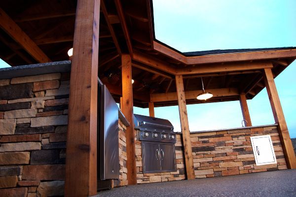 Covered outdoor patio with wood beams, stone wall, and a built-in grill under a blue sky