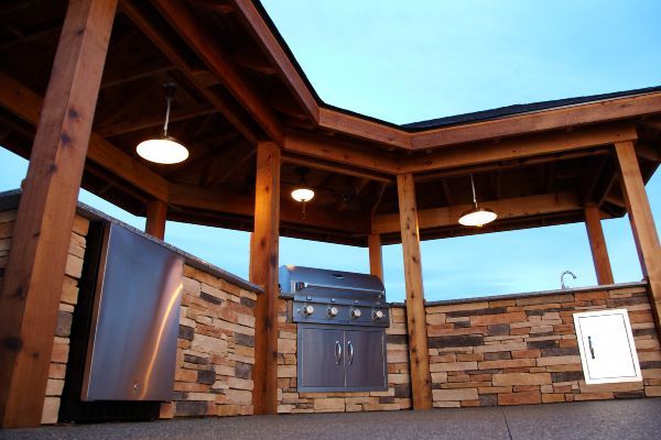 Outdoor patio kitchen with stainless grill, stone backsplash, wooden beams, and hanging lights under a blue sky