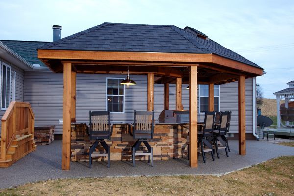 Wooden backyard gazebo with a dark shingled roof, chairs, and a hanging light on a patio at dusk