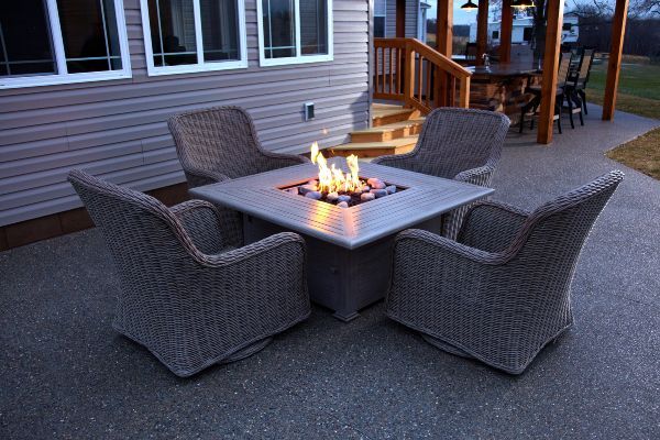 Outdoor patio seating with four wicker chairs around a square fire pit table with a lit flame.