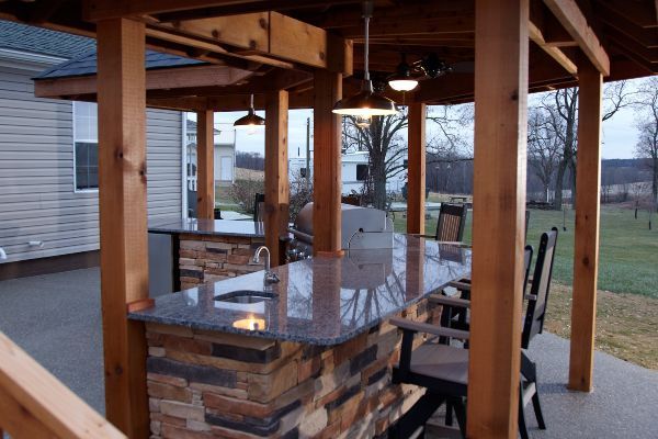 Covered outdoor patio with wooden posts, stone bar counter, and glass-top table overlooking a yard