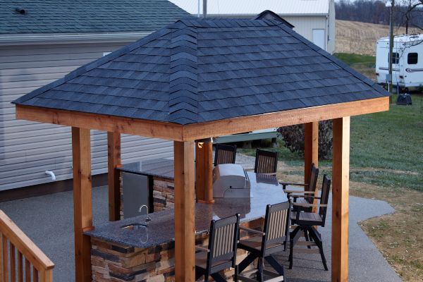 Wooden gazebo with shingled roof and patio furniture on a deck beside a house