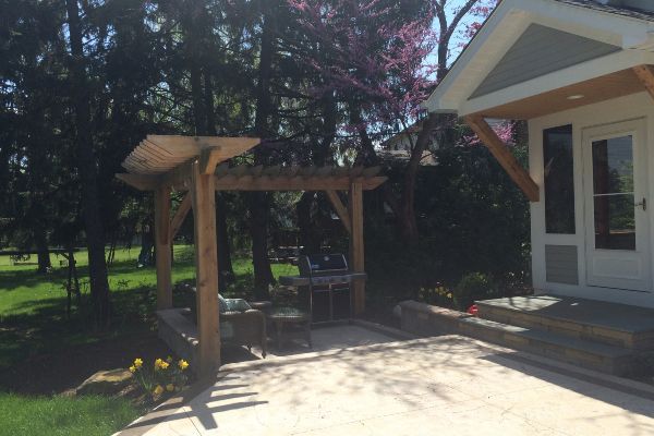 Backyard patio with wooden pergola, outdoor seating, and a house entrance beside a paved area