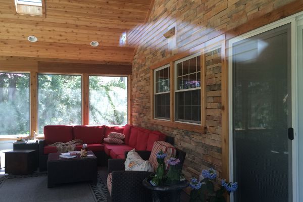 Covered sunroom with red sectional sofa, brick walls, wood ceiling, and large windows.