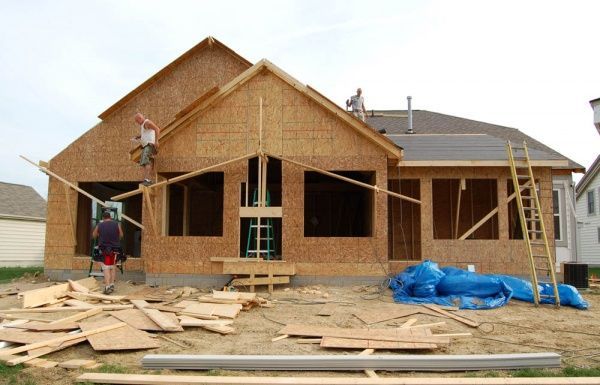 House under construction with unfinished wood framing, workers on scaffolding, and building materials scattered outside