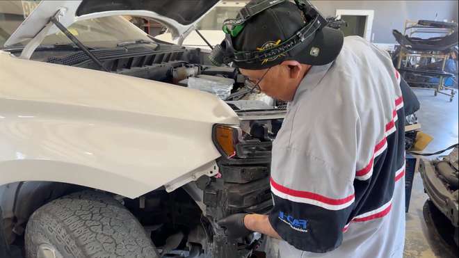 A mechanic wearing a headlamp works on the front engine area of a white vehicle in an auto repair shop.