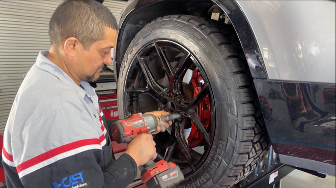 A technician uses a power tool to tighten lug nuts on a black car wheel with red brake calipers in an auto shop.