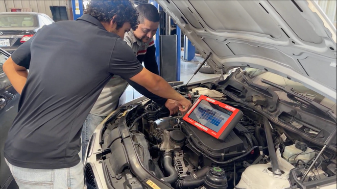 Two people in a garage work on an open car engine, using a diagnostic tablet to check the vehicle's computer system.