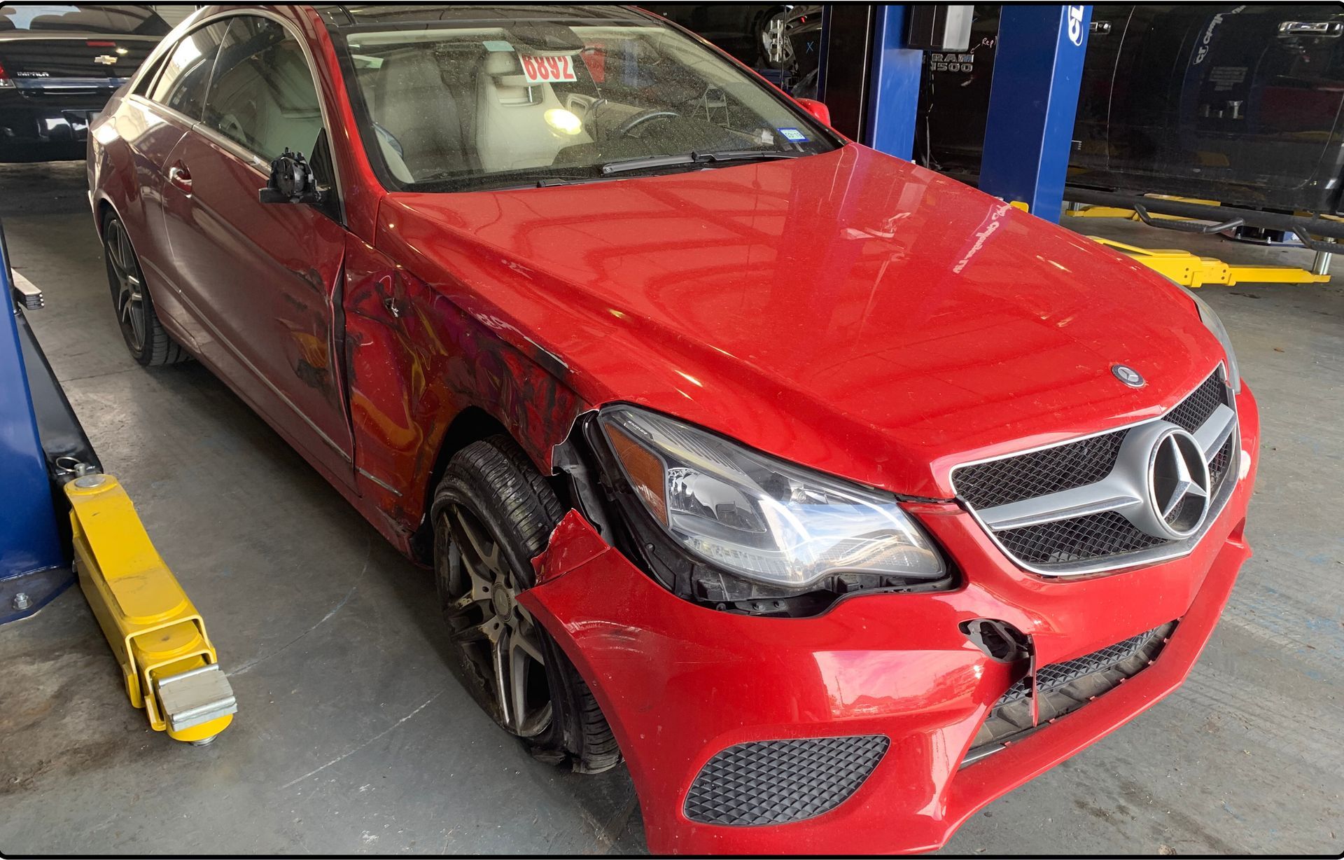 A red Mercedes-Benz coupe with front-end damage parked in an auto repair shop next to a vehicle lift.