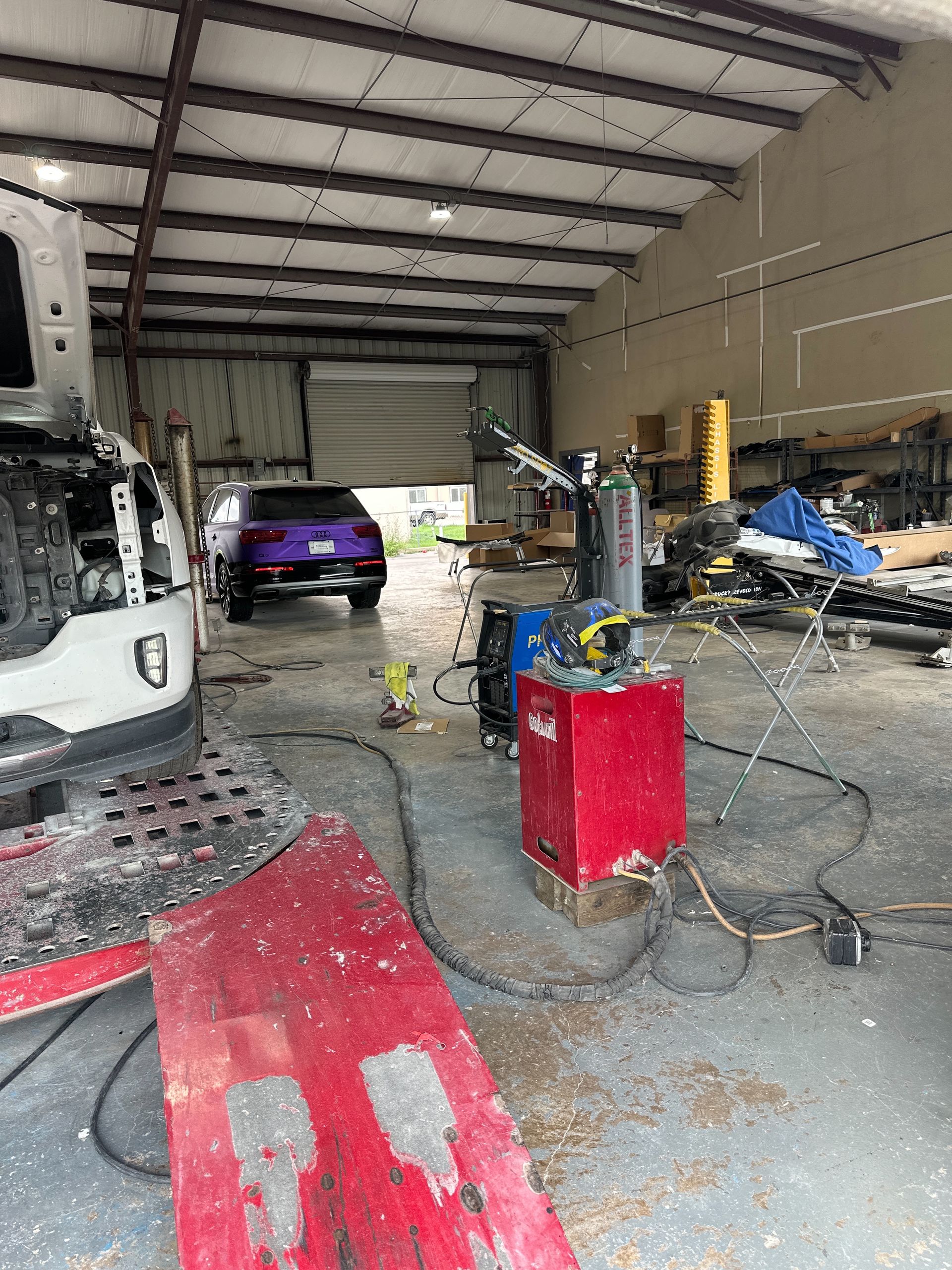 A cluttered auto repair shop with a white car on a lift, a purple car in the background, and red welding equipment.