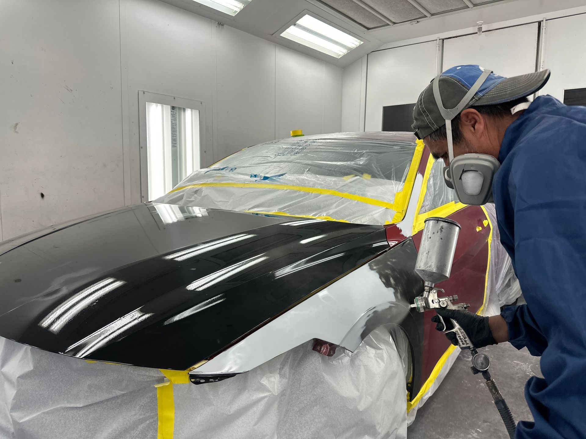 Person spray-painting a black car in a paint booth, wearing a respirator and protective gear.