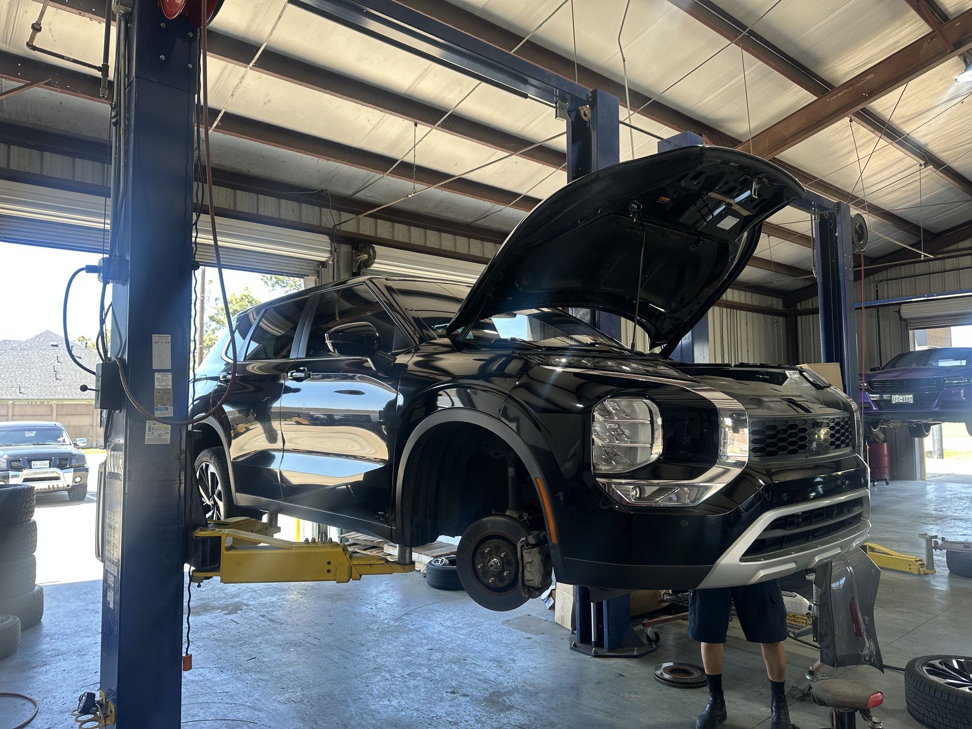 Black SUV lifted in auto shop with hood open, mechanic working underneath.