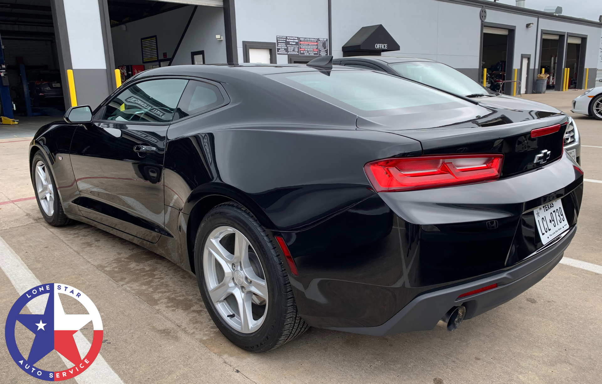A black Chevrolet Camaro parked outside a service center with a Texas-themed logo in the corner.