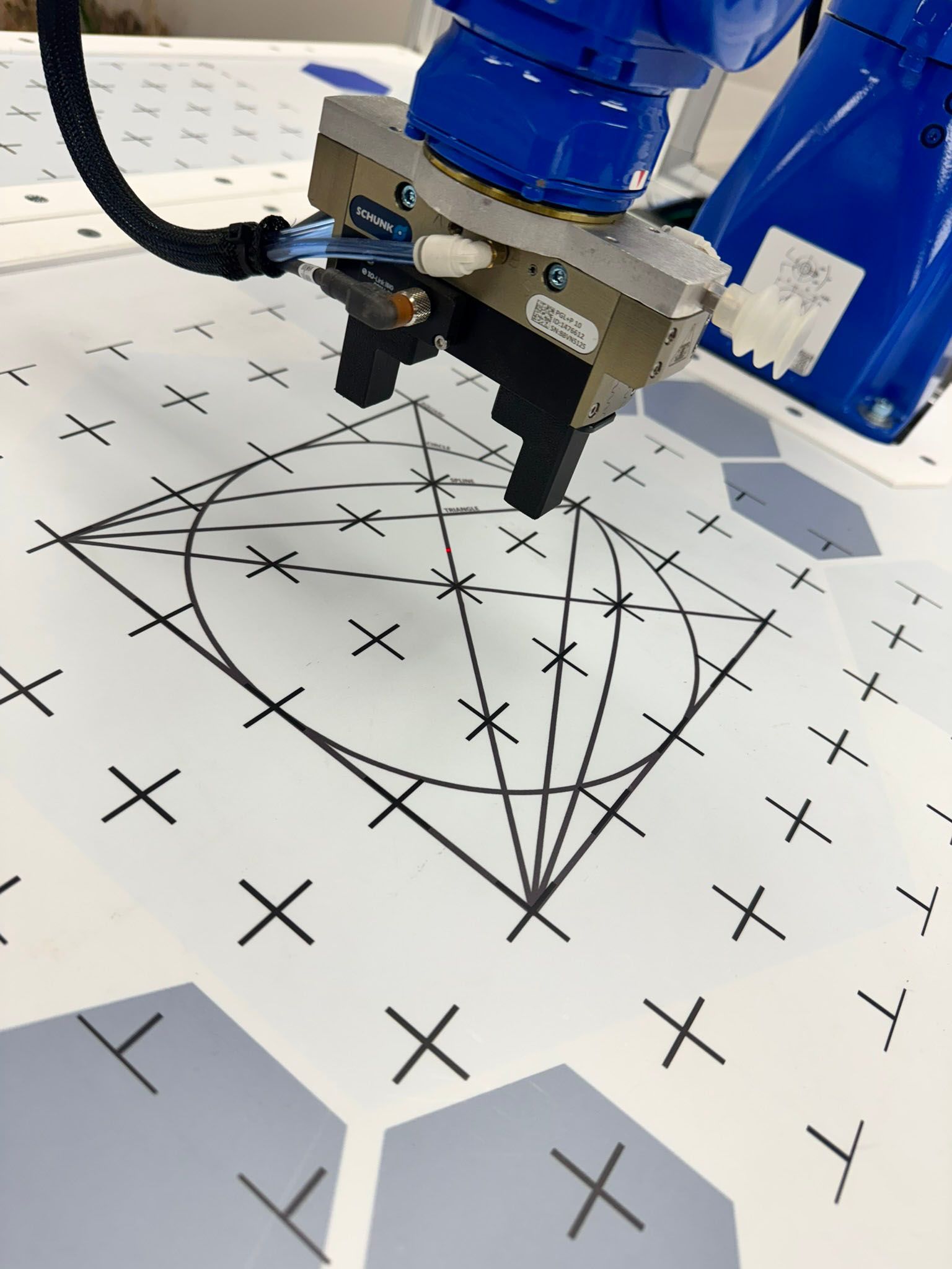 Robot arm drawing a geometric pattern on white paper with black grid marks.