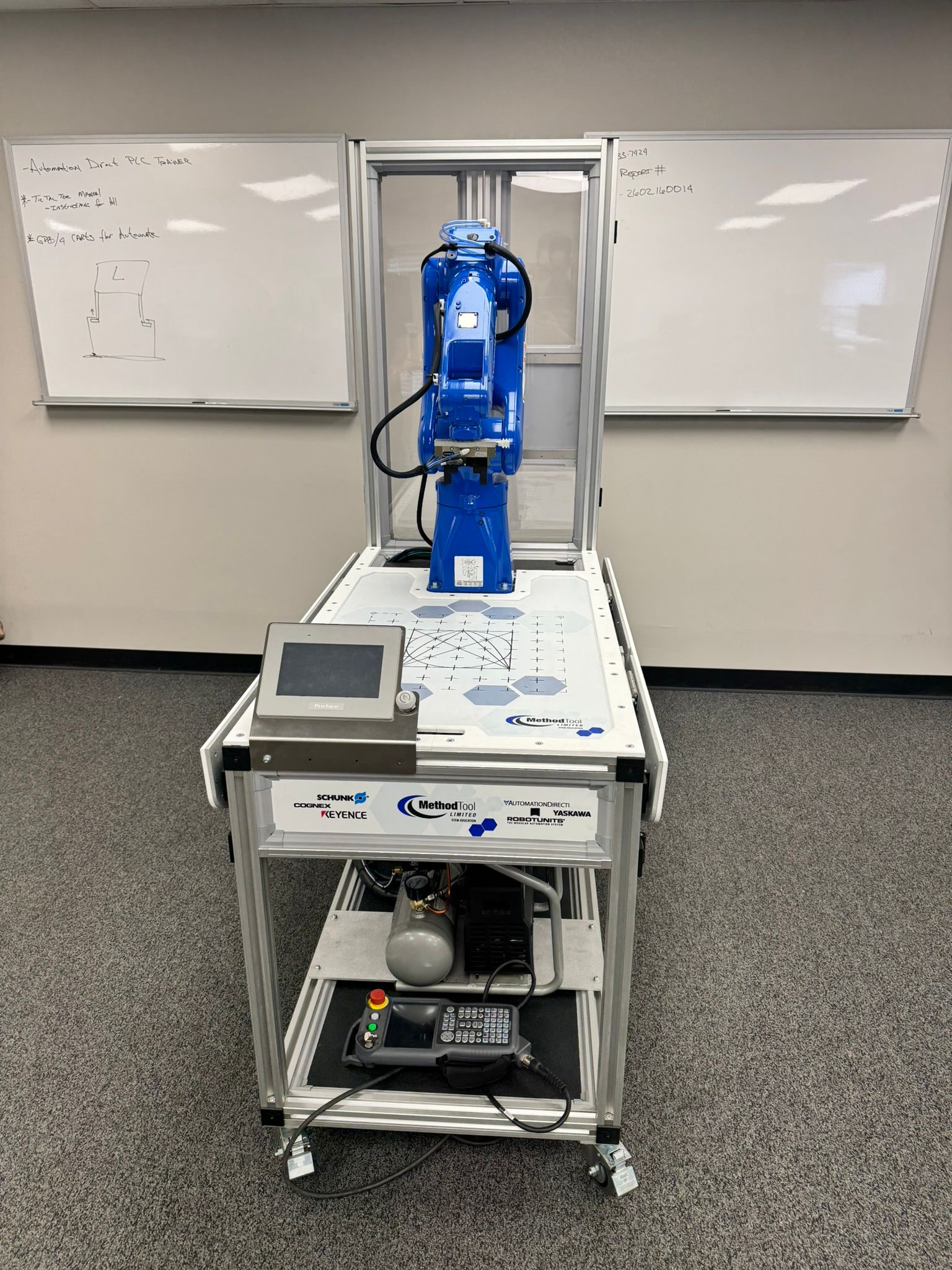 Blue industrial robot arm on a wheeled cart in a classroom or lab, with whiteboards in the background.