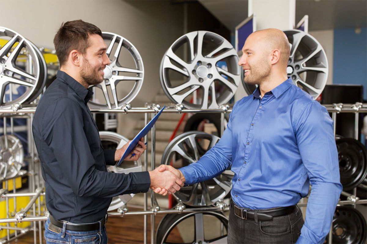 Two men shaking hands in a tire shop, with alloy wheels displayed in the background.