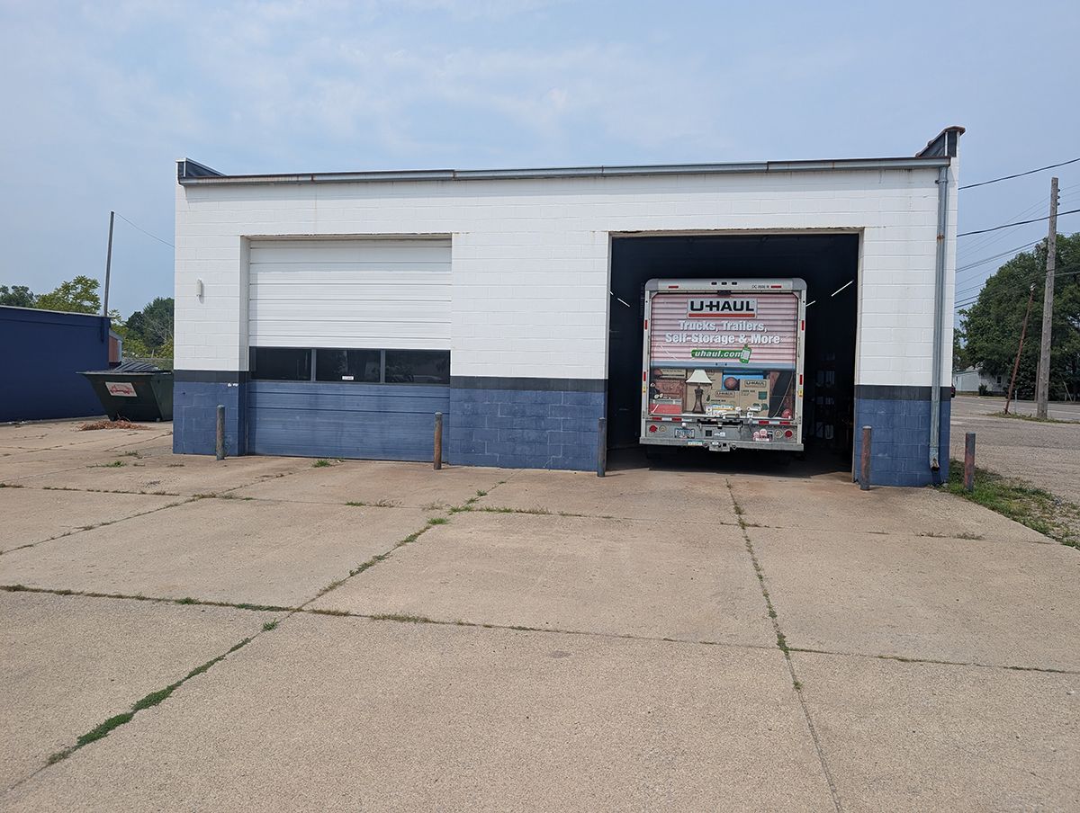 Small white-and-blue industrial building with a large open garage entrance and closed window under a cloudy sky
