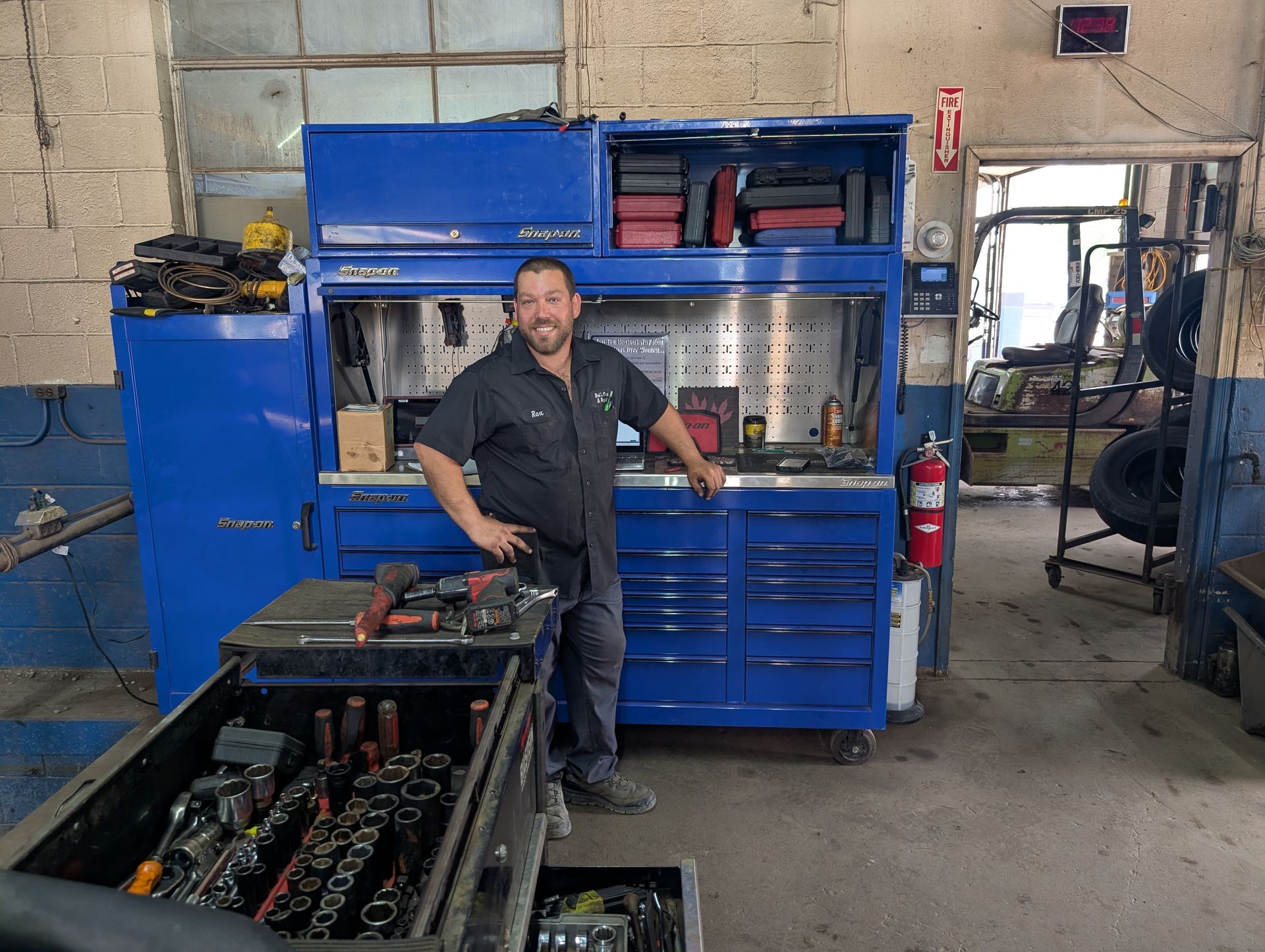 Person standing in a workshop beside a blue industrial machine and metal parts racks.