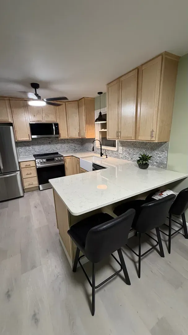 Modern kitchen with light wood cabinets, white island, black bar stools, and stainless steel appliances.