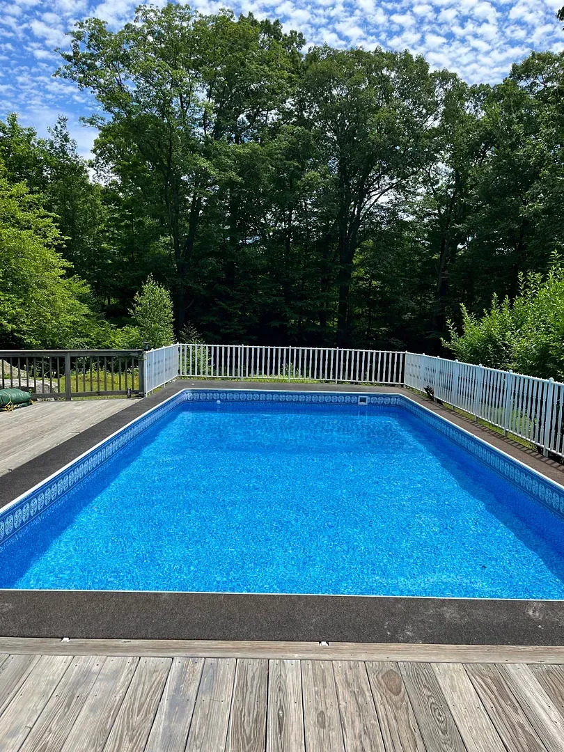 Backyard swimming pool with bright blue water, white fence, and trees under a partly cloudy sky