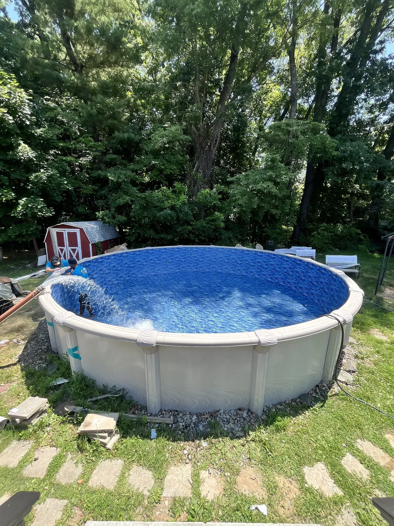 Above-ground pool filled with blue water in a grassy backyard, surrounded by trees and stepping stones