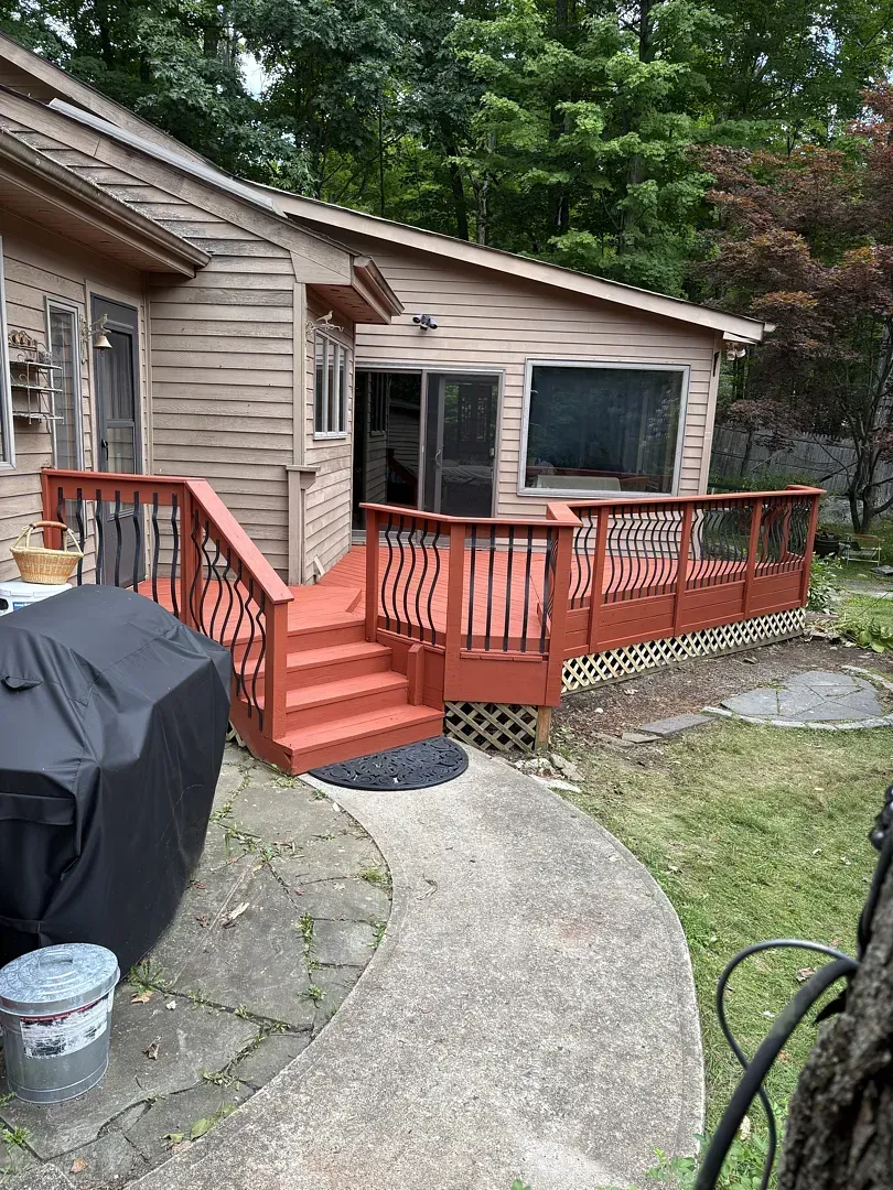 House with small red deck and stairs beside a curved concrete path, with trees in the background
