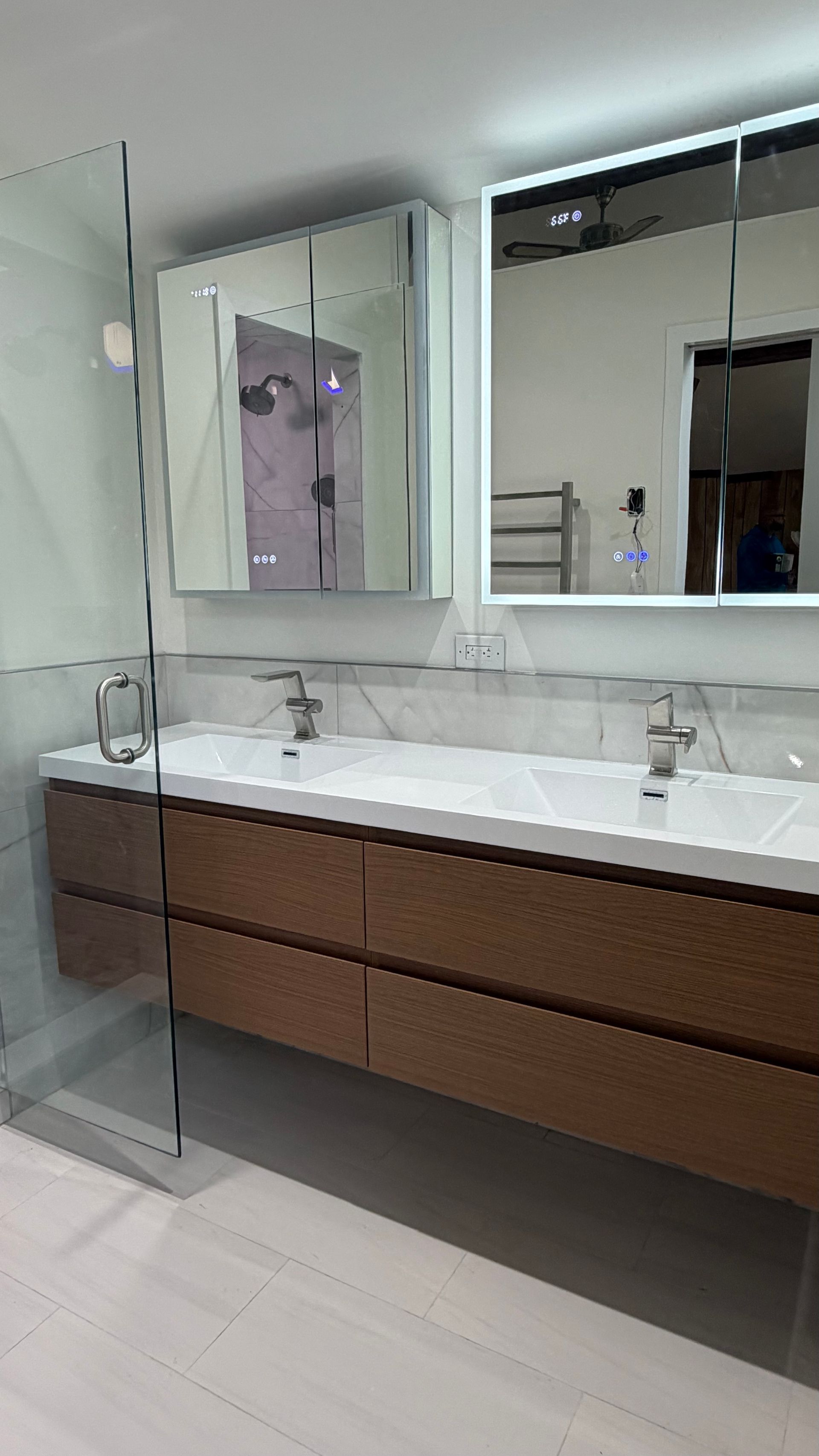Modern bathroom vanity with double mirrors, white countertop, and wood cabinets beside a glass shower.