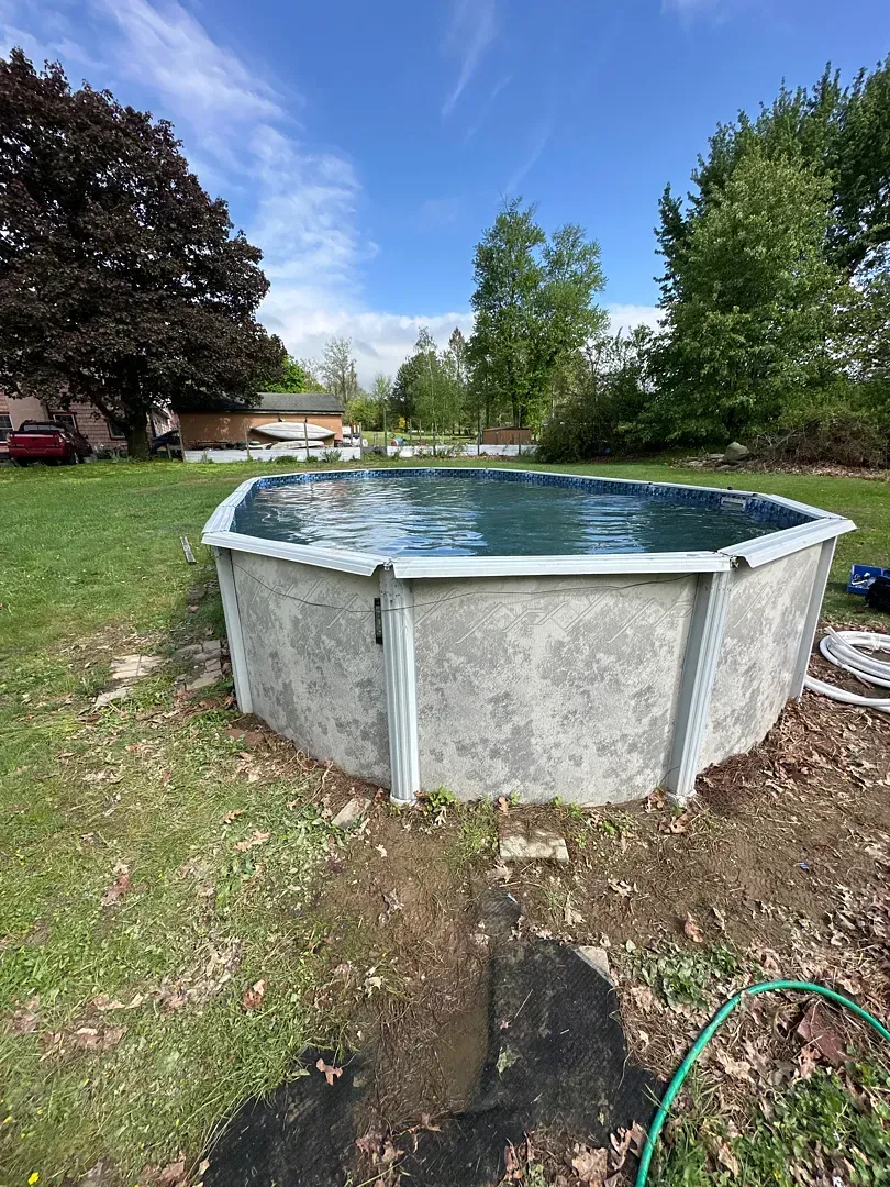 Above-ground round pool in a grassy backyard with trees and a house in the background.