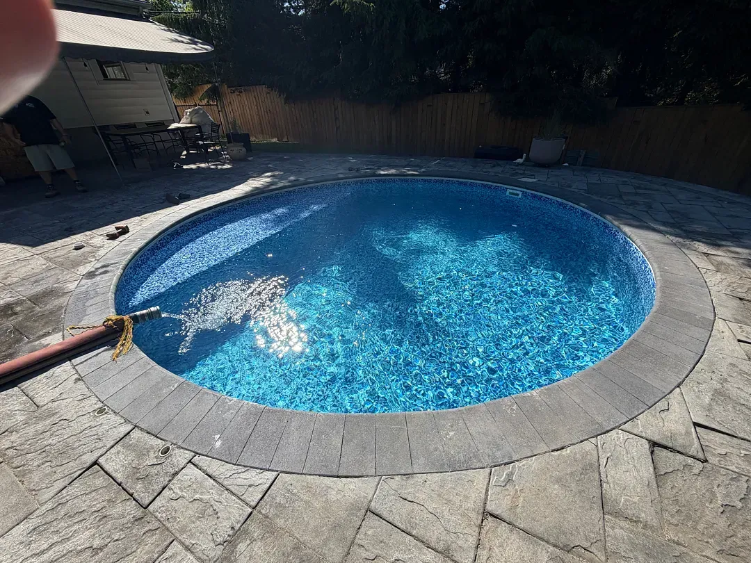 Backyard with a round blue swimming pool and stone patio, water splashing inside.