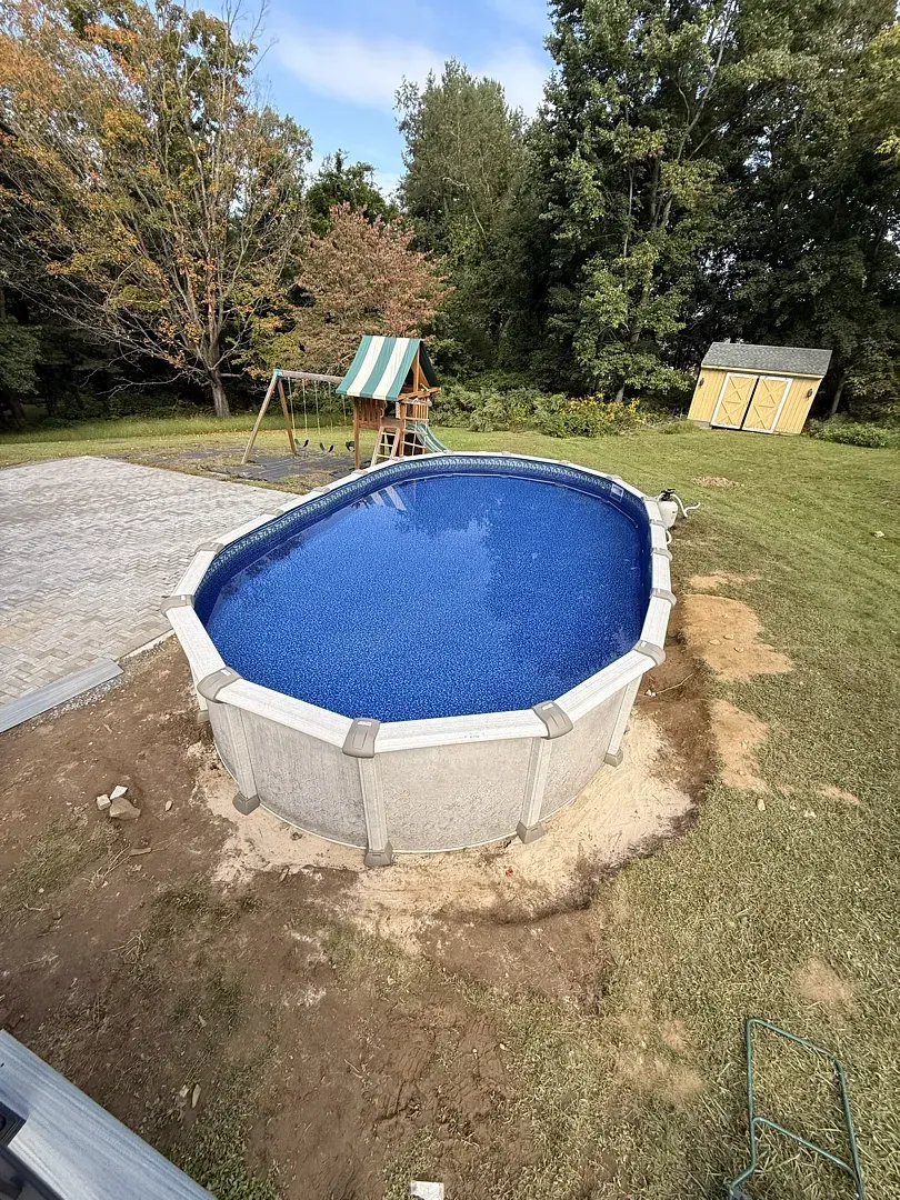 Above-ground round pool with blue cover in a grassy backyard, trees and two small sheds in the background