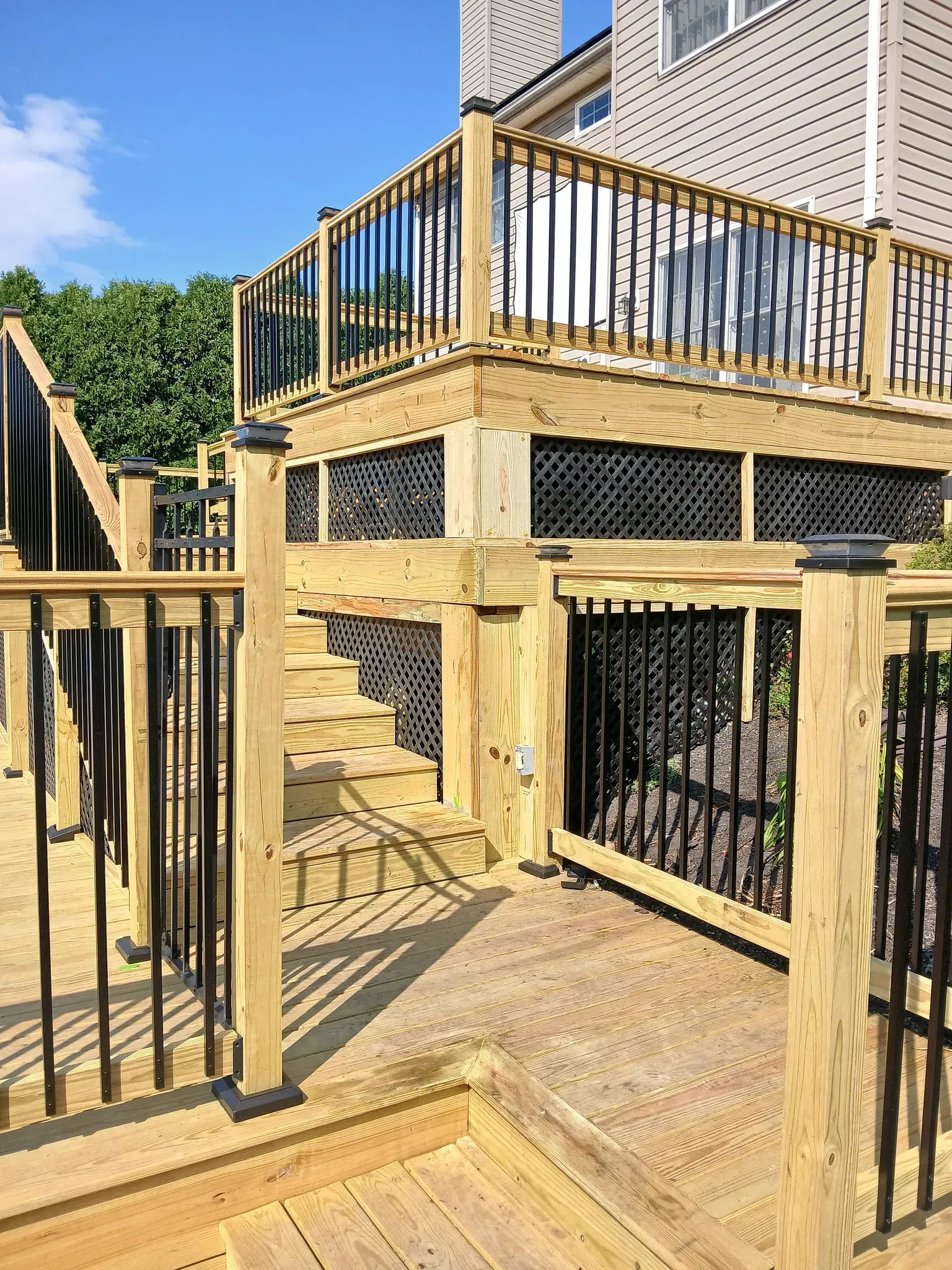 Wooden deck staircase and railing outside a house, with black metal gates and stone foundation.